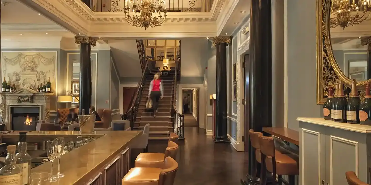 A bar with numerous stools and an elegant chandelier with person in red walking up staircase.