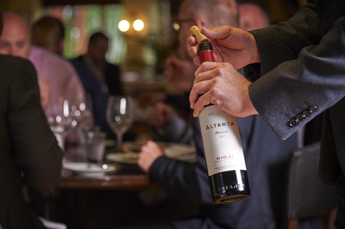 Sommelier opening a bottle of red Rioja wine for guests seated at a restaurant table, with wine glasses and warm ambient lighting.