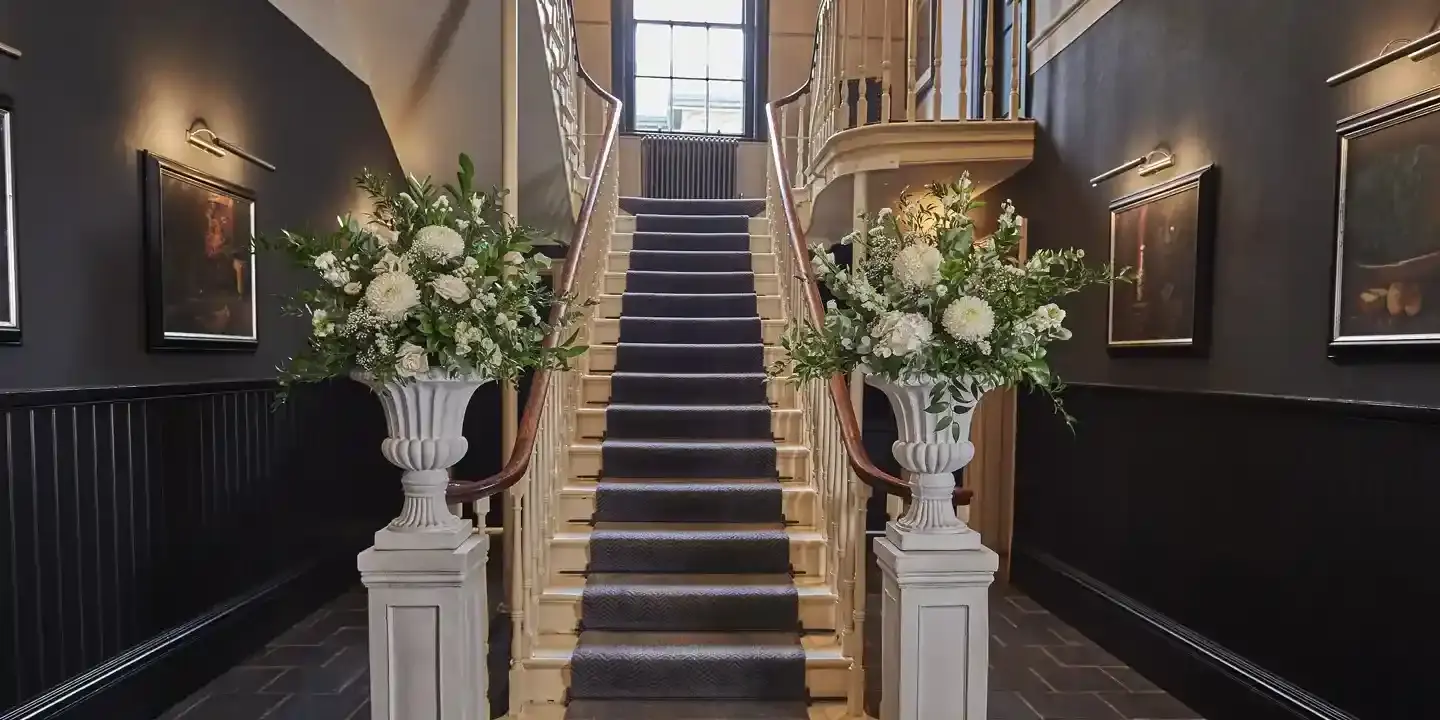 A hallway featuring a staircase and two vases filled with white flower bouquets.