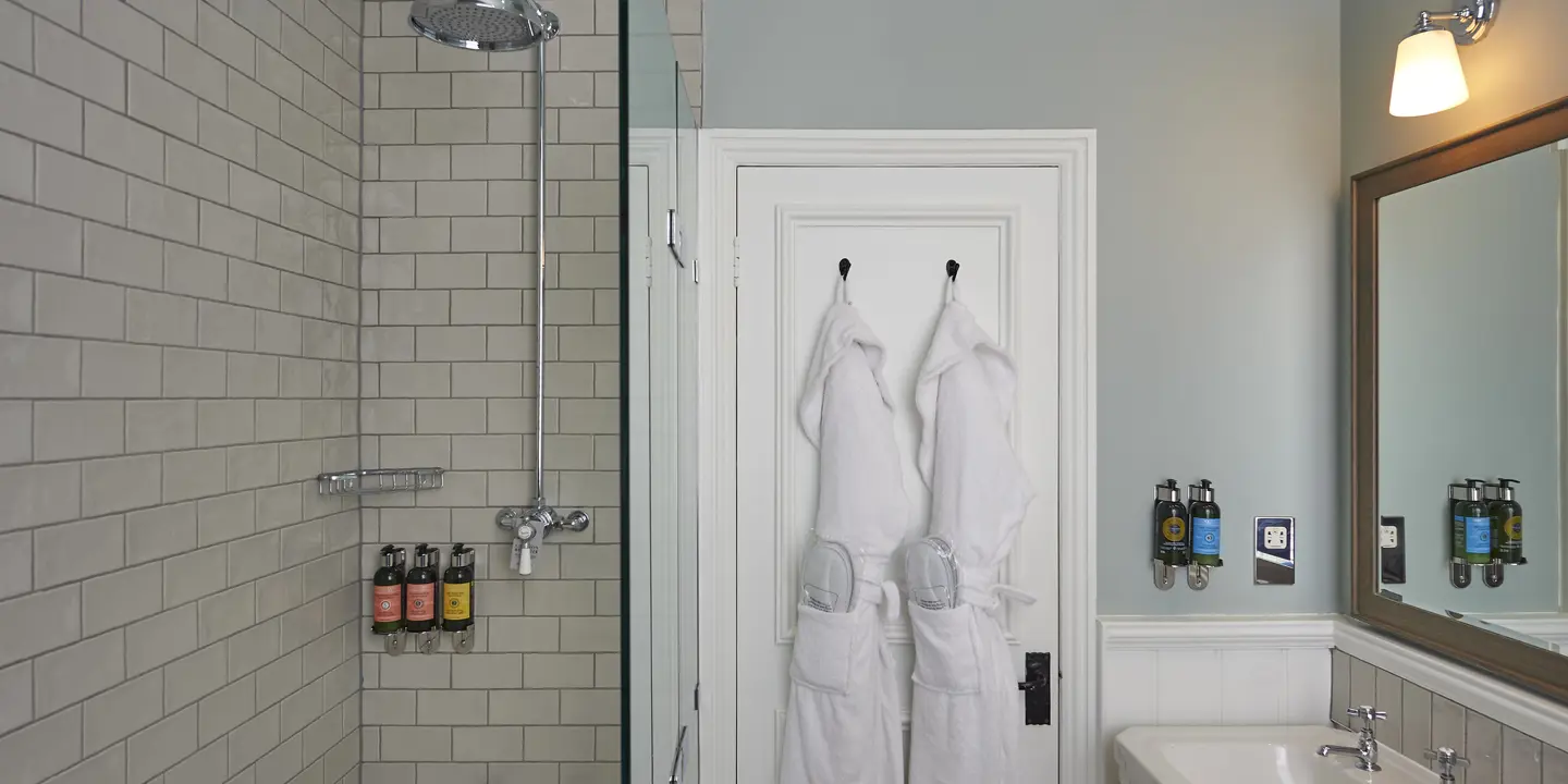 A bathroom featuring a sink, mirror, 2 dressing gowns hanging on the door and a bathtub.