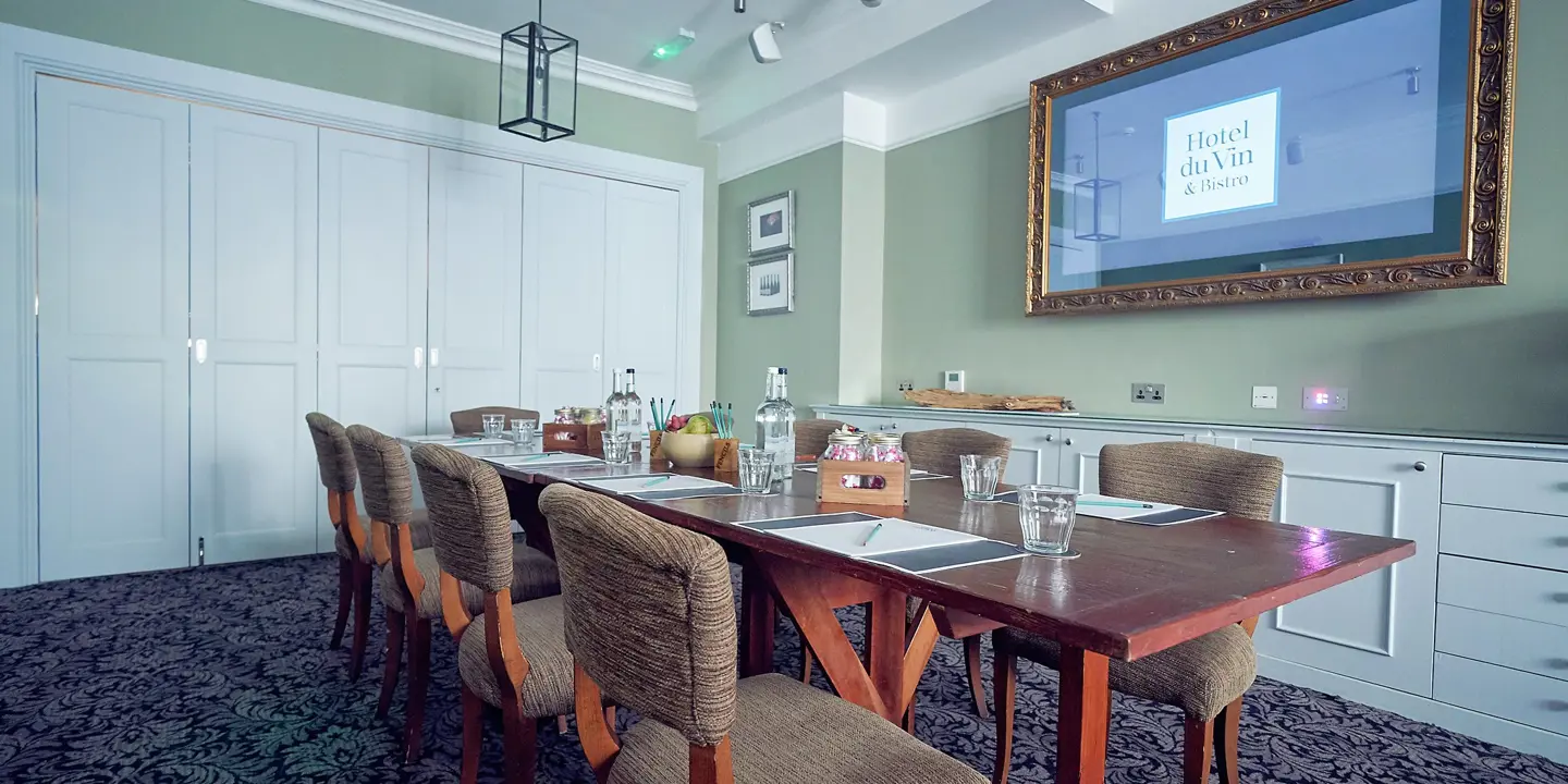 Conference room featuring a spacious wall-mounted screen, long table surrounded with chairs.
