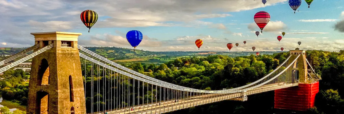 Numerous hot air balloons soar above a bridge.