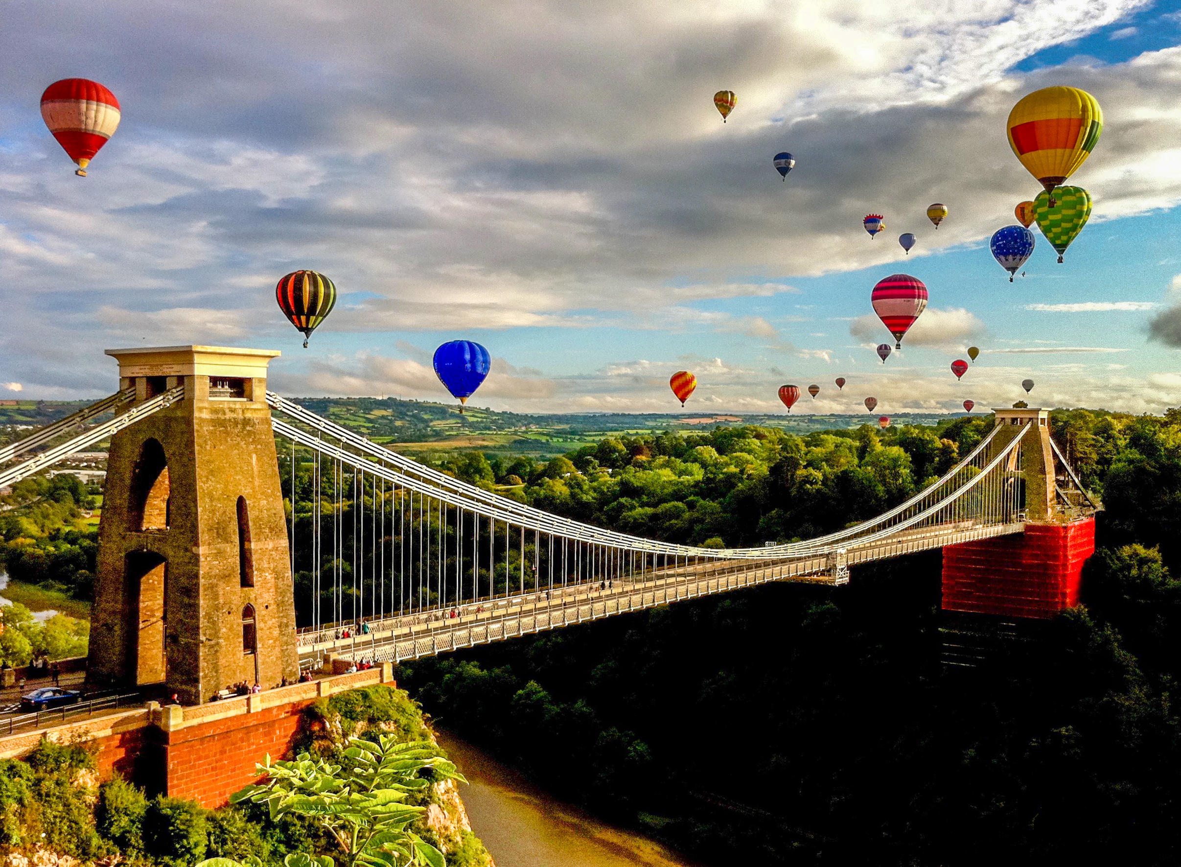 Numerous hot air balloons soar above a bridge.
