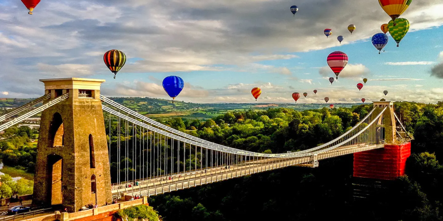 Numerous hot air balloons soar above a bridge.