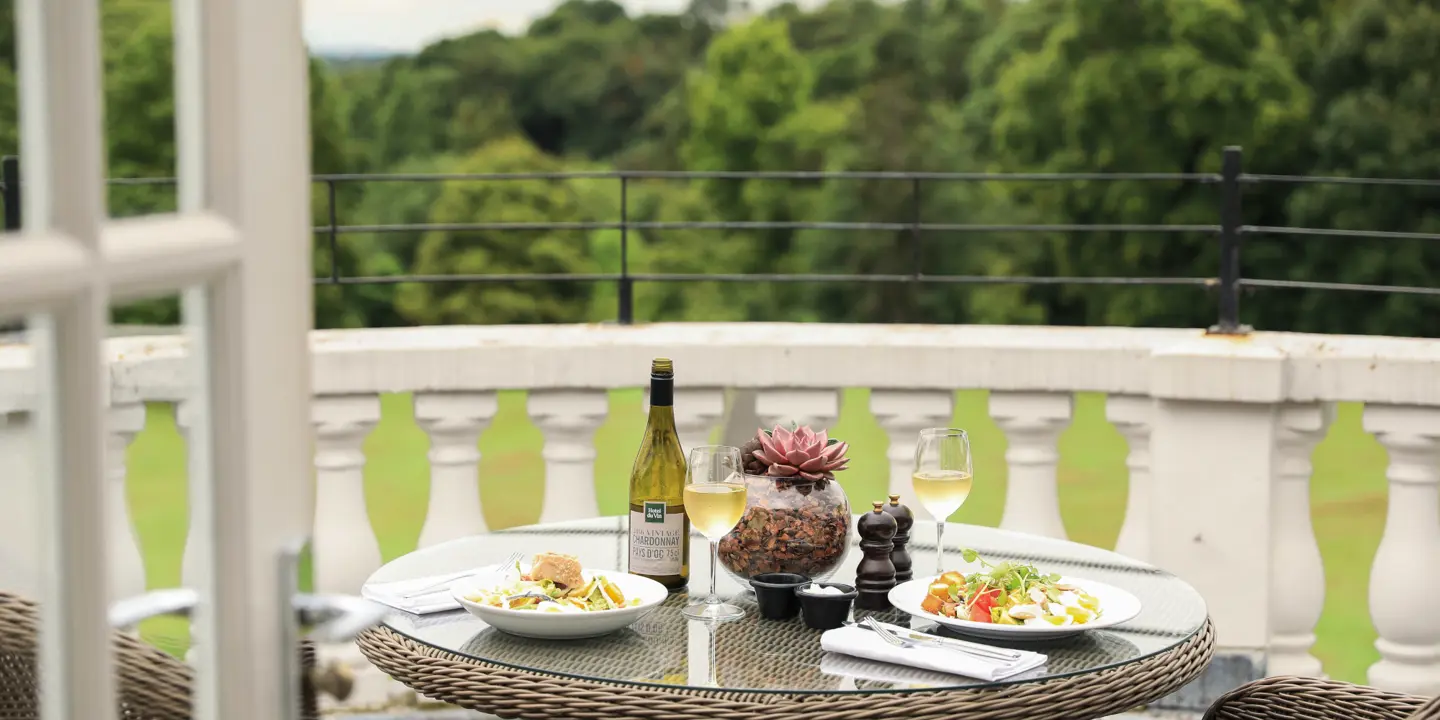 A table set on a balcony with two plates of food and a bottle of wine.