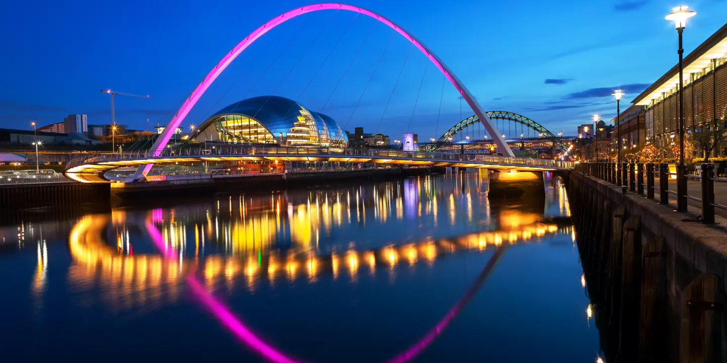 Night time view of the illuminated Millennium Bridge with The Glasshouse International Centre for Music in the background