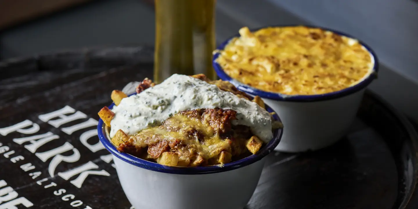 A close up of loaded fries, macaroni cheese, and a beer on a black tray