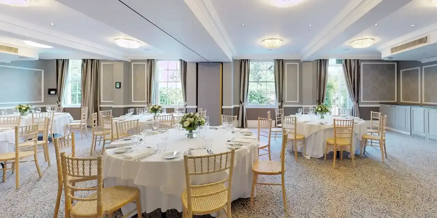 A spacious room adorned with neatly arranged round tables, chairs, and pristine white tablecloths.