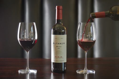 Red Rioja wine being poured into a glass beside a bottle of Altanza Reserva, with two wine glasses on a wooden table.