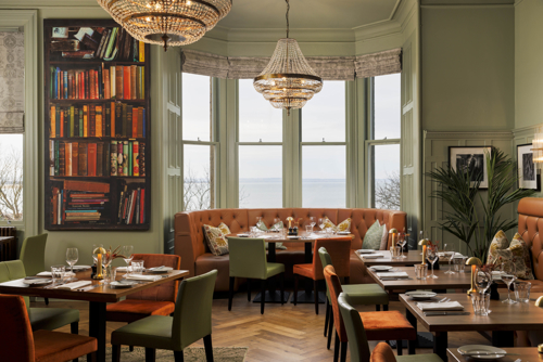 Elegant restaurant dining room with chandeliers, sage-green walls, orange banquette seating and neatly set tables overlooking the sea.