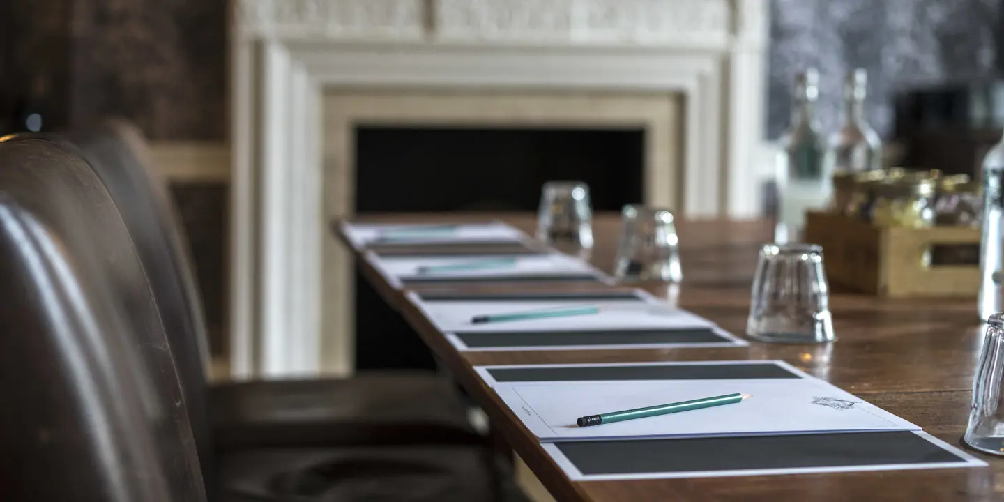 A close up of a wooden meeting room table, there is pen, paper, and empty water glasses visible 