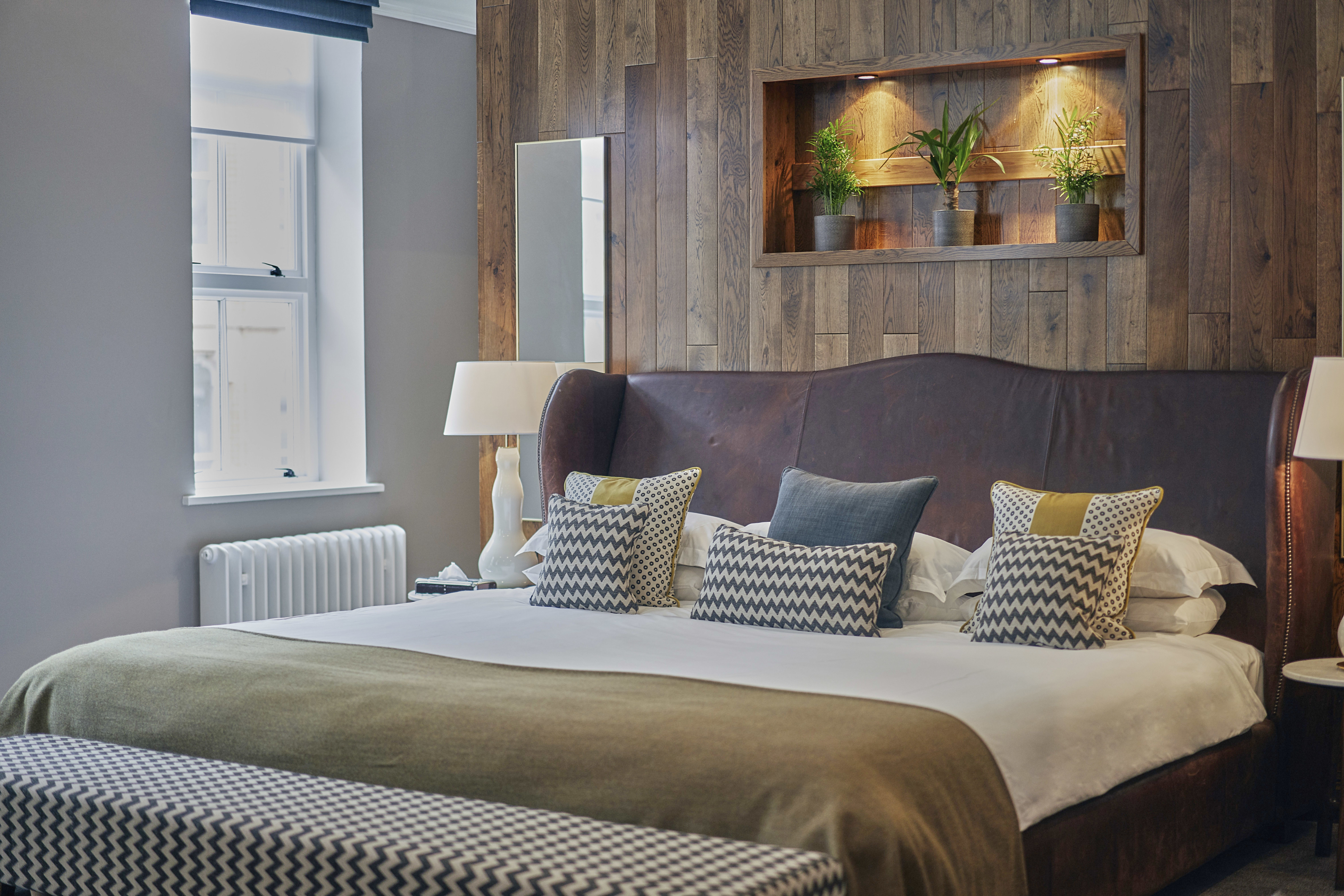 A dark brown leather double bed below a shelf of house plants. There is a dimly lit light shining on the shelf which holds the plants